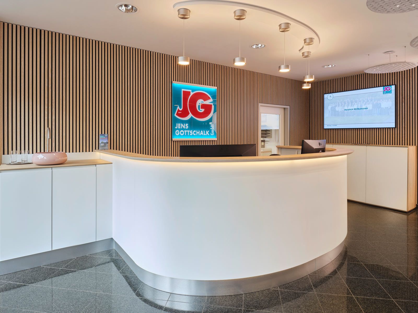 Empfangsbereich auf kleinem Raum - Die Verwendung von Echtholz-Akustikpaneelen verleiht dem Raum Wärme und eine klare Struktur. Die aluminiumfarbene Fußblende findet sich in den Holtkötter-Leuchten über dem Tresen wieder und rundet das Erscheinungsbild gekonnt ab.